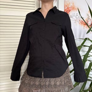 May include: Black long-sleeve button-down shirt with two chest pockets. The shirt has a classic collar and buttoned cuffs. The shirt is paired with a brown skirt.