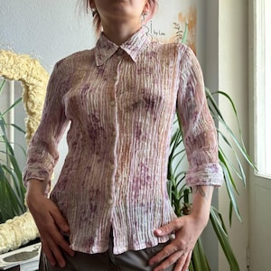 Puede incluir: Una camisa de manga larga con botones, textura arrugada y una base clara con rayas verticales y un suave estampado floral en tonos violeta y beige. La camisa cuenta con un cuello clásico, una tapeta de botones frontal y puños.