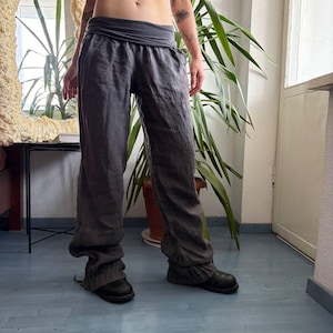 May include: Charcoal grey trousers featuring a wide, folded waistband and a relaxed, straight-leg fit. The trousers are constructed from a textured fabric and include gathered, elasticated cuffs at the ankles, providing a loose silhouette.