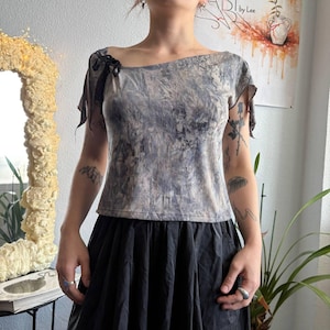 May include: A short-sleeved top featuring an asymmetrical neckline and a marbled gray and beige pattern. The garment includes a small black ribbon detail on the right shoulder and has a fitted silhouette that ends at the waistline.