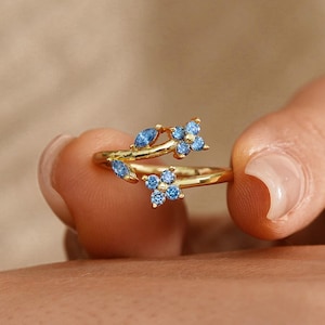 Può includere: Un anello dorato con un design floreale, ornato da piccole pietre preziose blu. L'anello è tenuto tra le dita, mettendo in risalto i suoi delicati dettagli e la sua maestria artigianale. Le pietre preziose sono disposte per assomigliare a fiori.