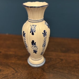May include: A white ceramic vase with blue floral designs. The vase has a fluted body, a flared rim, and a round base. Blue trim accents the rim and base. The vase is approximately 10 inches tall.