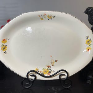 May include: Cream-colored oval serving platter with yellow floral accents and a black trim. The vintage dish is displayed on a black metal stand. The flowers are yellow, orange, and red.