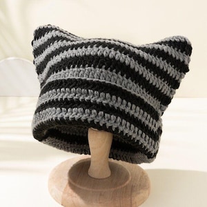 May include: A striped crochet hat in black and gray, featuring pointed ear-like details at the top. The hat is displayed on a wooden stand, showcasing its handmade texture and design. This unique accessory adds a playful touch to any outfit.