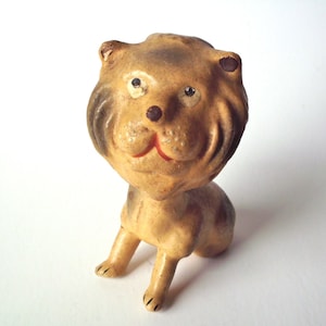 May include: A vintage lion figurine with a tan body and a brown mane. The lion has a smiling red mouth, black eyes, and is posed in a seated position. The figurine is made of a ceramic material.