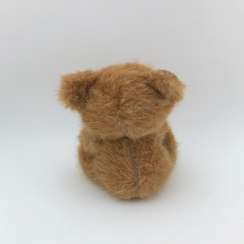 May include: A small, plush teddy bear in a light brown color. The bear is viewed from the back, showing its fluffy fur and the shape of its head and body. The toy is set against a plain white background.