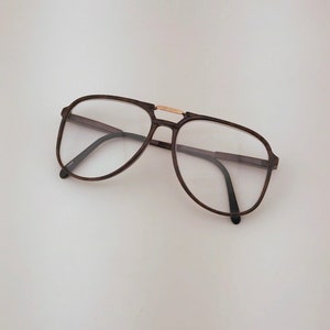 May include: Brown aviator-style eyeglasses with clear lenses. The frames have a dark brown color with a gold-colored bridge. The glasses are set against a white background.