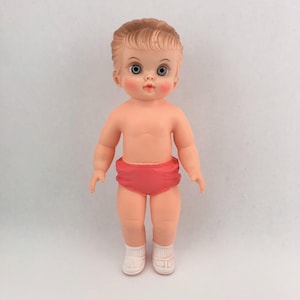May include: A plastic doll with light skin, molded brown hair, and large blue eyes. The doll features rosy cheeks and a small mouth, wearing bright pink briefs and white molded sandals. The figure stands in an upright position with arms at its sides.