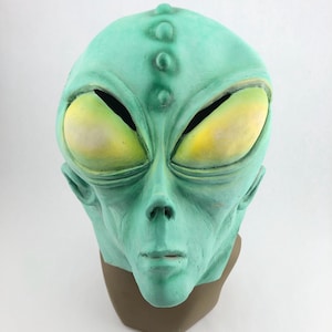 May include: A green alien mask with large yellow eyes and a textured surface. The mask has a prominent forehead and a detailed facial structure, including a nose, mouth, and ears. The mask is made of a durable material and is suitable for costume parties or Halloween.