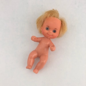May include: A small, vintage plastic baby doll with a peach skin tone, blue eyes, and blonde hair. The doll is nude and in a playful pose, with arms outstretched and legs bent. The doll is a classic toy.