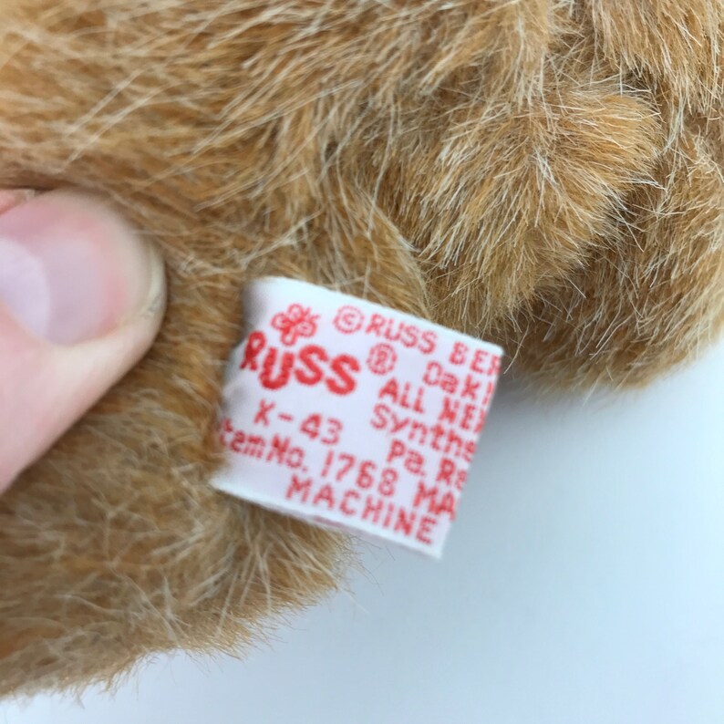 May include: Close-up of a plush toy's fur with a white tag. The tag has red text that reads "RUSS BER" and other details. The fur is light brown and fluffy.