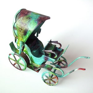 May include: A miniature, decorative carriage with a canopy, painted in shades of green, teal, and maroon. The carriage features spoked wheels and curved handles, suggesting a vintage or antique style. The overall design is whimsical and artistic.