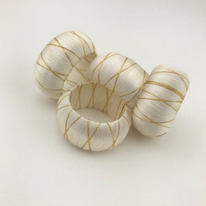 May include: A collection of four ivory-colored napkin rings. Each ring is wrapped in a silky material and accented with a gold crisscross pattern. The rings are arranged on a white surface, showcasing their elegant design.
