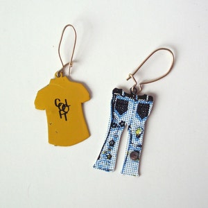 May include: A pair of novelty earrings. One earring is a yellow t-shirt shape with black text. The other earring is a blue and white denim jeans shape with black details and a button.