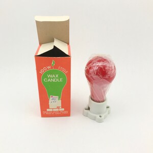 May include: A red wax candle shaped like a lightbulb, wrapped in clear plastic, sits in a white porcelain socket. Beside it is an orange box with the words "Wax Candle" and "100W" printed on it.