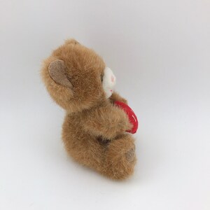 May include: A small, plush teddy bear in shades of brown, holding a red heart. The bear has a white muzzle and pink accents on its nose and mouth. The heart has a white stitched design.