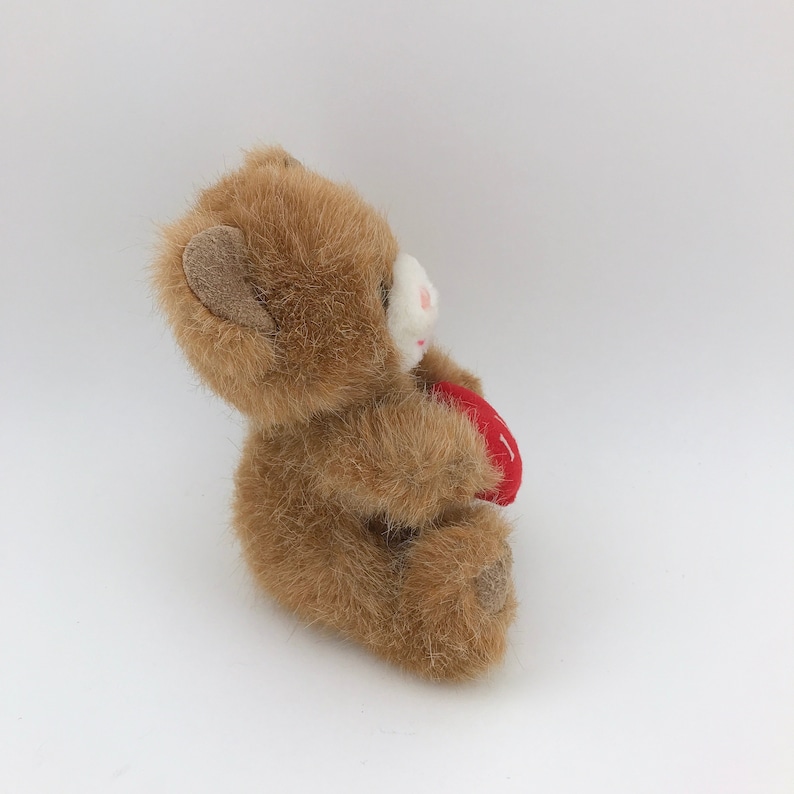 May include: A small, plush teddy bear in shades of brown, holding a red heart. The bear has a white muzzle and pink accents on its nose and mouth. The heart has a white stitched design.