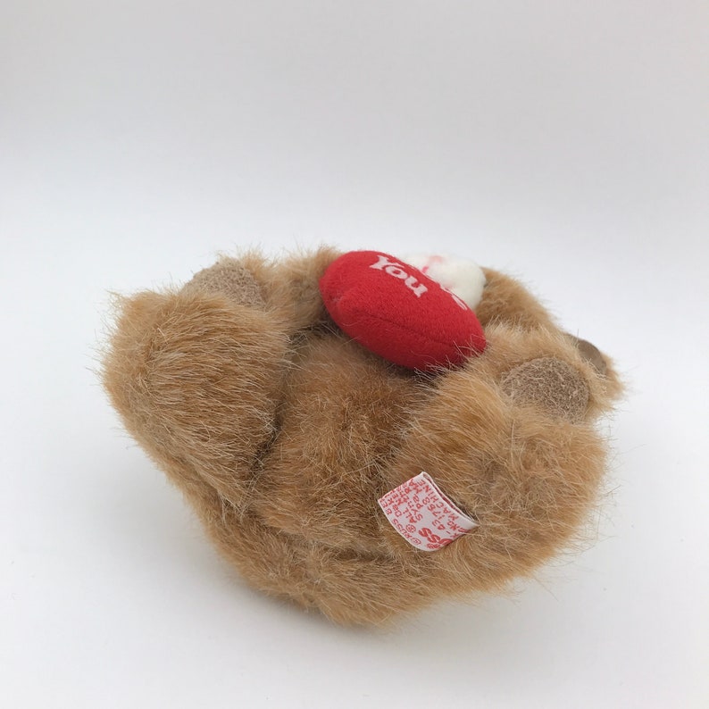 May include: A small, brown plush bear with a red heart-shaped pillow that says "You". The bear is lying down, with its paws visible. The plush toy has a tag attached.