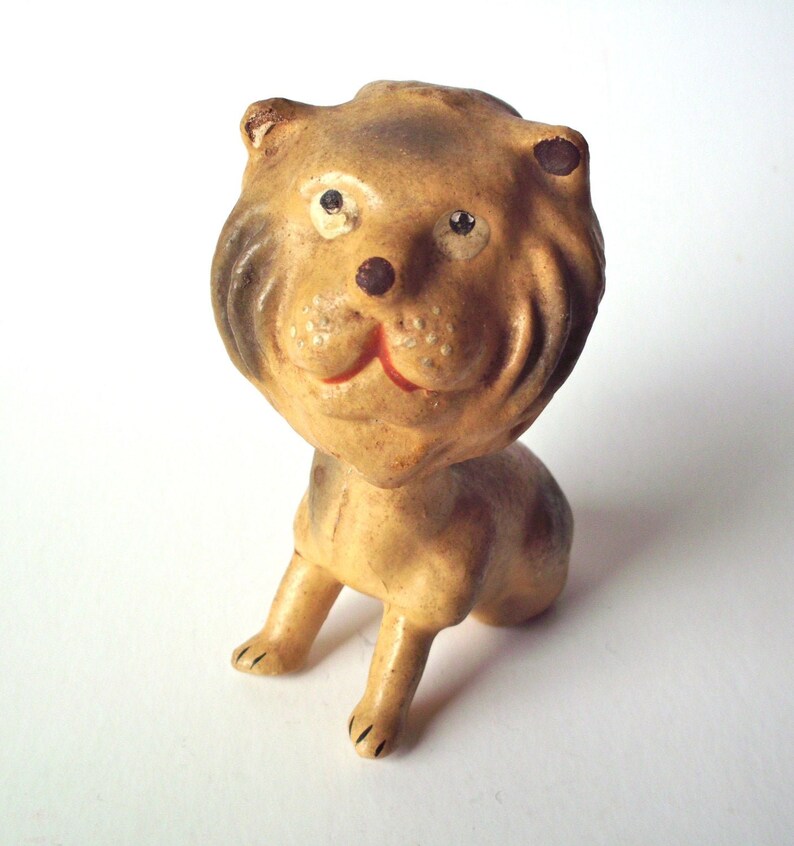 May include: A vintage lion figurine with a tan body and a brown mane. The lion has a smiling red mouth, black eyes, and is posed in a seated position. The figurine is made of a ceramic material.