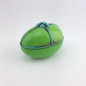 May include: A green, egg-shaped trinket box with turquoise accents. The box features a hinged lid and is adorned with turquoise beads and trim. The overall design is a vibrant combination of colors and textures, suitable for storing small items.