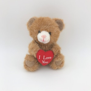 May include: A small brown teddy bear with a white muzzle and a pink nose. The bear is holding a red heart-shaped sign that reads "I Love You" in white letters. The bear has a soft, plush appearance.