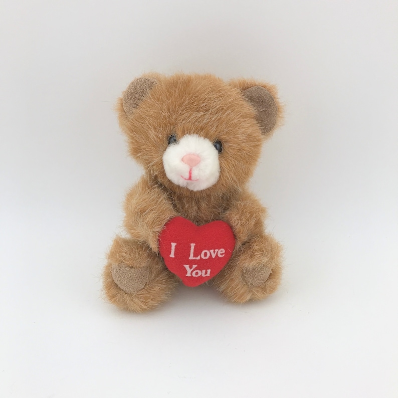 May include: A small brown teddy bear with a white muzzle and a pink nose. The bear is holding a red heart-shaped sign that reads "I Love You" in white letters. The bear has a soft, plush appearance.