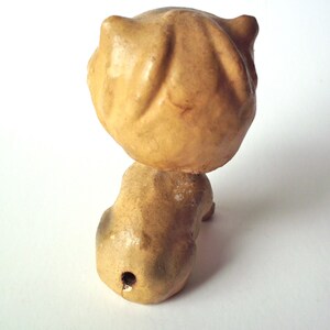 May include: A vintage, tan-colored figurine of a cat, viewed from the back. The cat's head is round with two small, pointed ears. The body is elongated, with a small hole near the base. The figurine has a distressed, aged appearance.