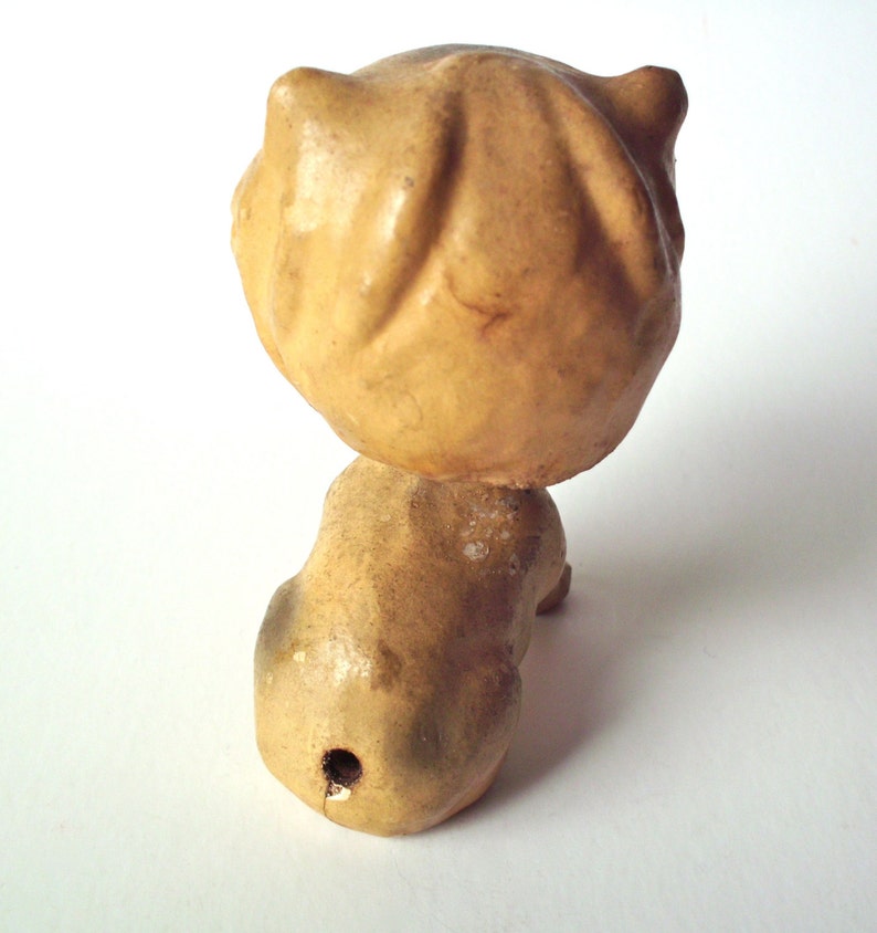 May include: A vintage, tan-colored figurine of a cat, viewed from the back. The cat's head is round with two small, pointed ears. The body is elongated, with a small hole near the base. The figurine has a distressed, aged appearance.