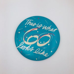 May include: A round, teal-colored decorative plate with the text "This is what 60 looks like" in white and pink script. The number 60 is in a cream color with a pink outline. Small pink and white accents are scattered around the plate.