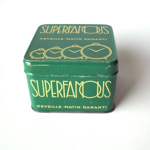 May include: A vintage green metal box with the words "SUPERFAMOUS" in gold lettering. The box also features the text "REVEILLE-MATIN GARANTI" and a series of gold alarm clock illustrations. The box is square-shaped.