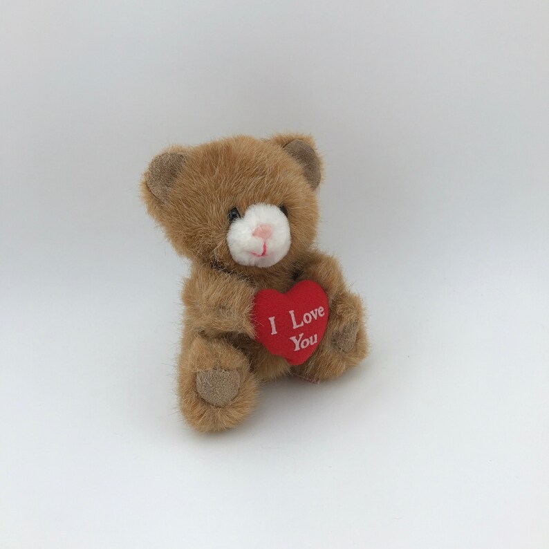 May include: A small, brown teddy bear holding a red heart. The heart has the words "I Love You" printed on it in white. The bear has a white muzzle and a pink nose. The plush toy is set against a white background.