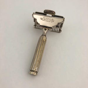 May include: A vintage silver-toned safety razor with the word "GEM" engraved on the head. The handle has a textured, ridged design for grip. The razor is designed for shaving.