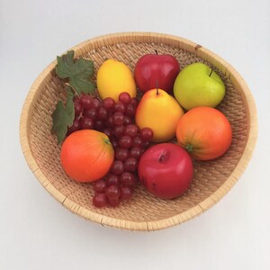May include: A round, light-tan woven wicker basket containing an assortment of artificial fruit, including a yellow lemon, a red apple, a green apple, a yellow pear, an orange, a red apple, and a cluster of red grapes with green leaves.