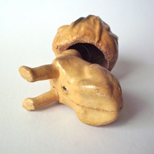 May include: A vintage, tan-colored ceramic figurine, possibly a toy or decorative item, with a bulbous head and a body with two legs. The head is open, revealing a dark interior. The figurine is lying on a white surface.