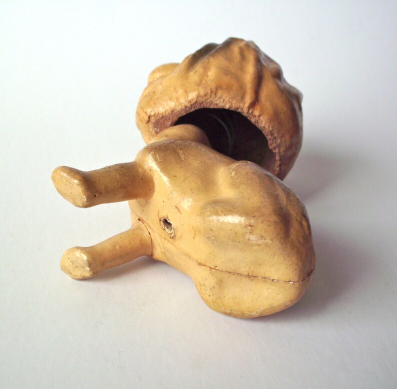 May include: A vintage, tan-colored ceramic figurine, possibly a toy or decorative item, with a bulbous head and a body with two legs. The head is open, revealing a dark interior. The figurine is lying on a white surface.