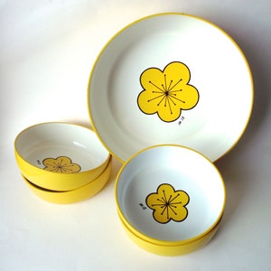May include: A set of white and yellow ceramic dishes. The set includes a large plate and several bowls. Each piece features a yellow flower design with black outlines. The rims and sides of the dishes are yellow.