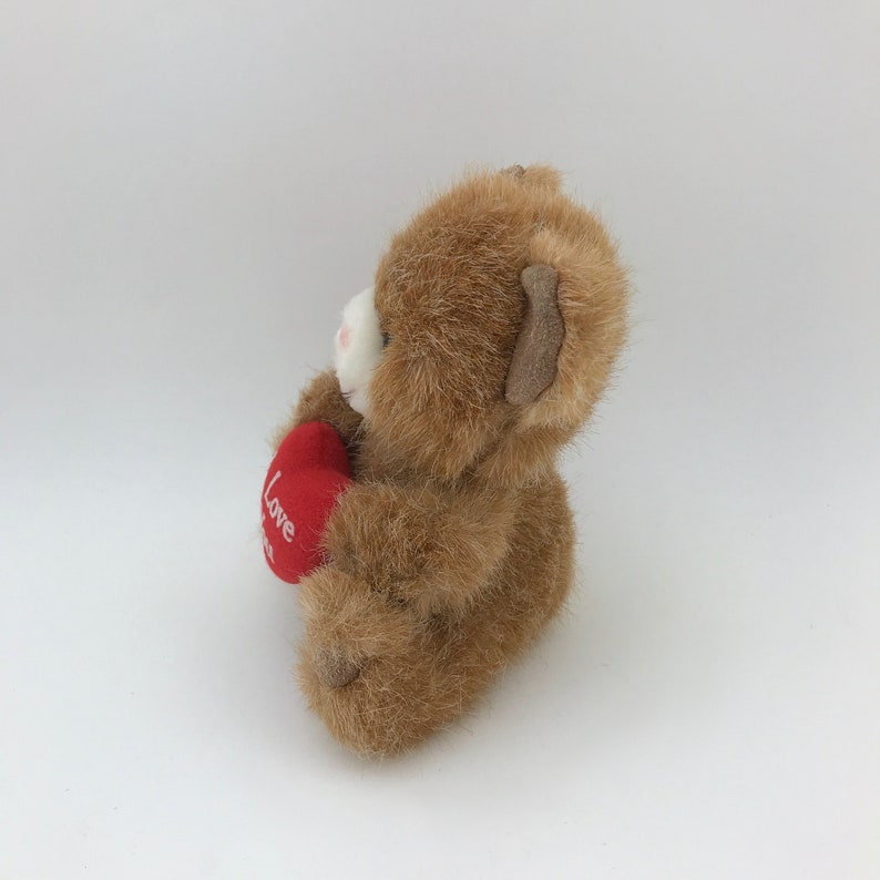 May include: A brown teddy bear holding a red heart with the word "Love" embroidered on it. The bear has a white muzzle and a light brown ear. The plush toy is set against a white background.