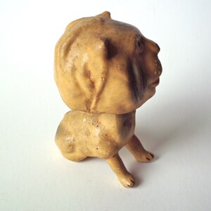 May include: A vintage, tan-colored figurine of a dog with a rounded head and body. The dog is depicted in a sitting position with its front legs extended. The figurine has a weathered appearance, suggesting it is an antique or a well-loved item.