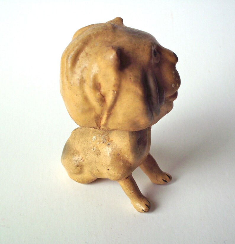 May include: A vintage, tan-colored figurine of a dog with a rounded head and body. The dog is depicted in a sitting position with its front legs extended. The figurine has a weathered appearance, suggesting it is an antique or a well-loved item.