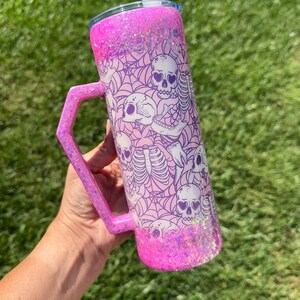 May include: A pink glitter tumbler with a handle, featuring a spooky design of skulls and spiderwebs. The tumbler has a lid and a black and white striped straw. The handle and bottom of the tumbler are covered in glitter.