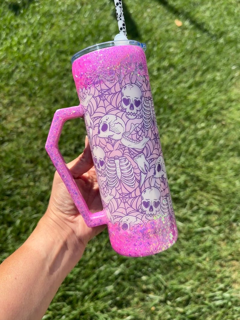 May include: A pink glitter tumbler with a handle, featuring a spooky design of skulls and spiderwebs. The tumbler has a lid and a black and white striped straw. The handle and bottom of the tumbler are covered in glitter.
