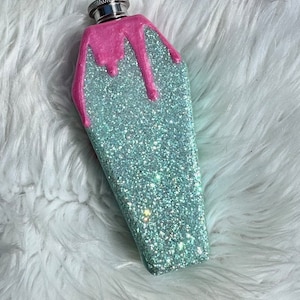 May include: A coffin-shaped flask with a silver cap. The flask is covered in sparkling, light blue glitter. Pink glitter drips down from the top of the flask, creating a unique design. The flask is a novelty item.