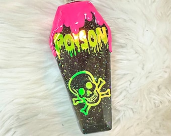 May include: A black coffin-shaped flask with a silver cap. It features a neon pink top and the word "POISON" in dripping green letters. A neon green skull and crossbones graphic is on the front.