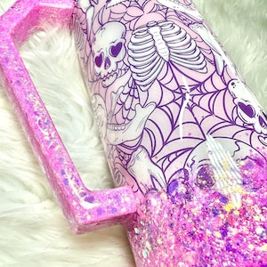 May include: A pink glitter tumbler with a matching handle. The tumbler features a spooky design with skulls, rib cages, and spiderwebs in white and purple against a light pink background. The bottom half is covered in pink and purple glitter.