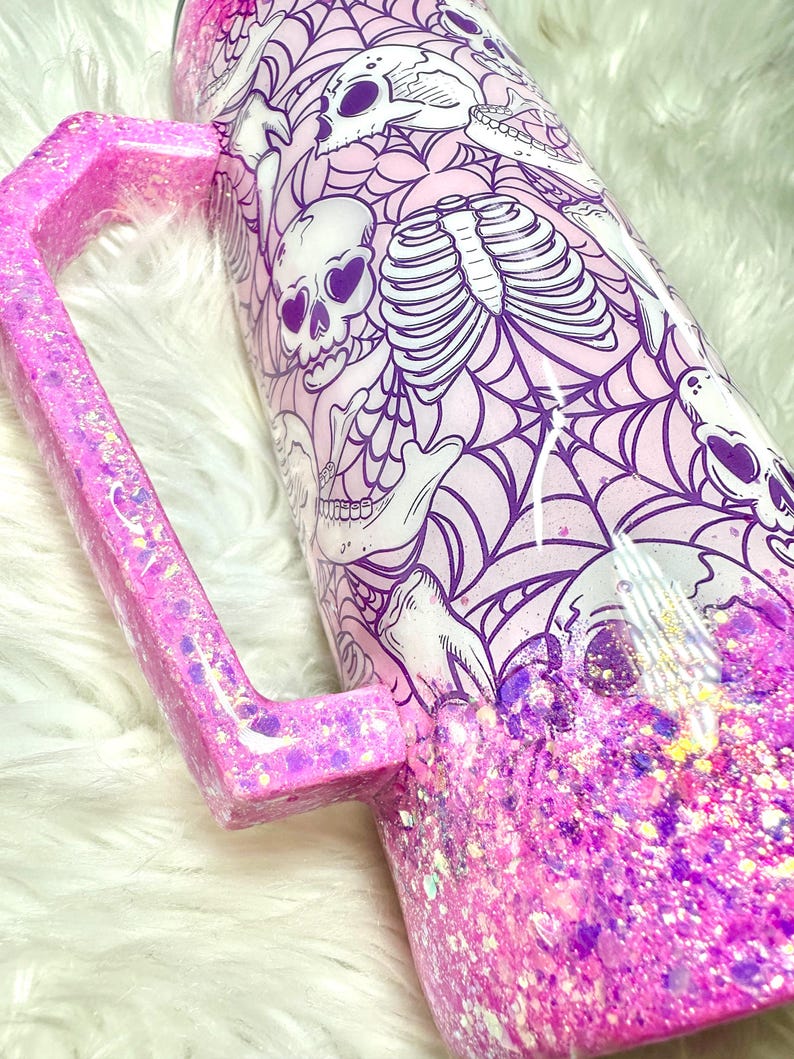 May include: A pink glitter tumbler with a matching handle. The tumbler features a spooky design with skulls, rib cages, and spiderwebs in white and purple against a light pink background. The bottom half is covered in pink and purple glitter.