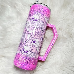 May include: A pink glitter tumbler with a handle, featuring a repeating pattern of purple skulls, spiderwebs, and skeletons. The tumbler has a black and white striped straw and a matching glitter handle. The bottom of the tumbler is covered in pink glitter.