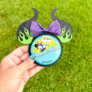 Pode incluir: Orelhas de Minnie Mouse verdes e pretas com um laço de purpurina roxo e um broche redondo que diz "Feliz aniversário!" com uma imagem do Mickey Mouse usando um chapéu de festa e um bolo com velas. O broche também diz "Disneyland" na parte inferior.
