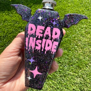 Puede incluir: Una petaca negra con forma de ataúd con purpurina y texto rosa que gotea que dice "DEAD INSIDE". La petaca tiene alas plateadas en el lateral.