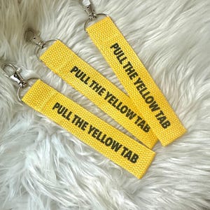 Op de afbeelding: Drie gele sleutelhangers met zilveren sluitingen. Elke sleutelhanger heeft de tekst "PULL THE YELLOW TAB" in zwarte letters. De sleutelhangers zijn gemaakt van geweven stof en worden gepresenteerd op een wit, pluizig oppervlak.
