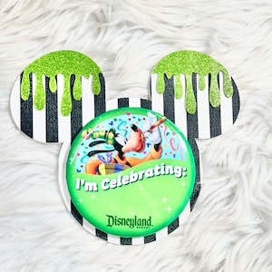 May include: Green glitter Mickey Mouse ears with a black and white striped background. The center features a round button with a cartoon image of Goofy celebrating with a party hat, confetti, and a gift. The button reads "I'm Celebrating: Disneyland Resort".